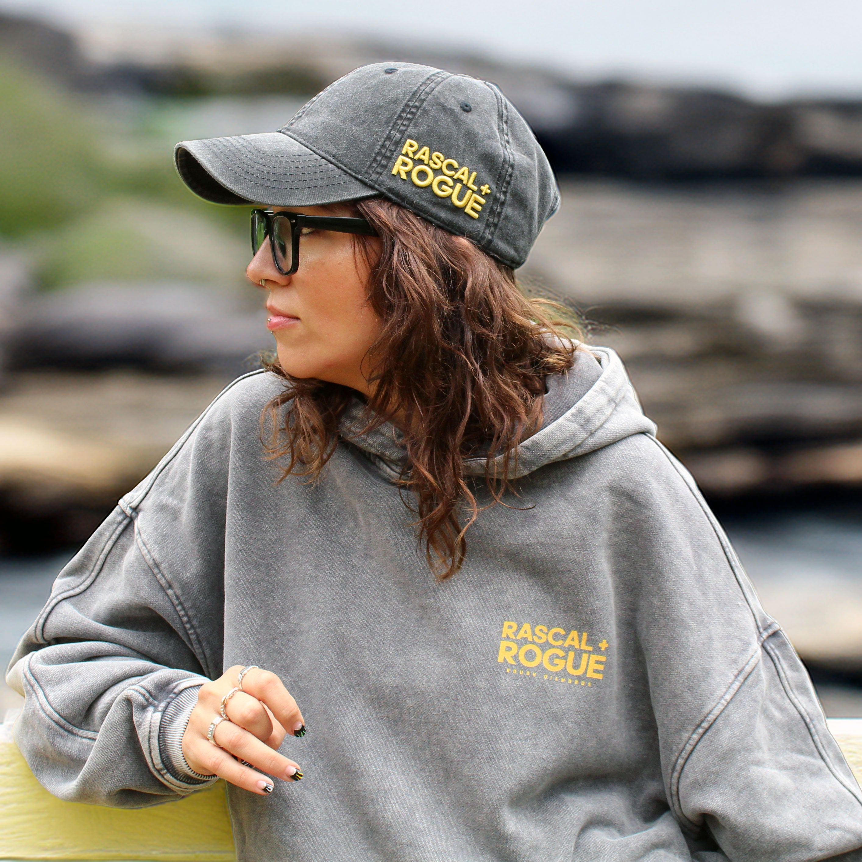 Person wearing a gray 'Rascal & Rogue' hoodie and cap outdoors.